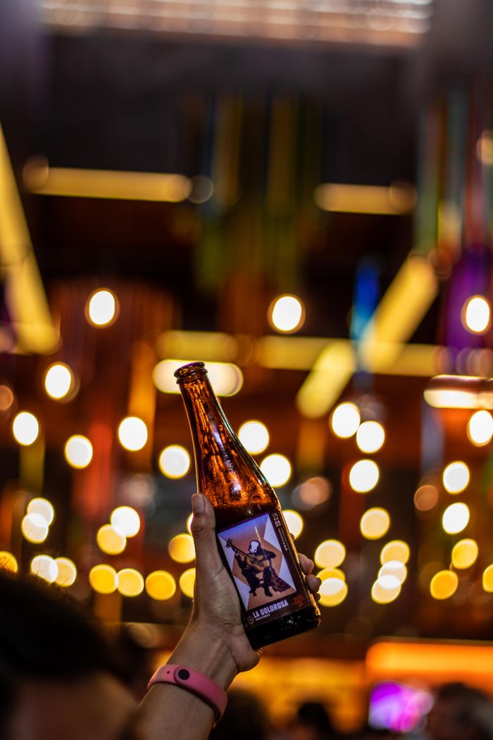 A hand holds a beer bottle against a backdrop of colorful bokeh lights indoors at night.