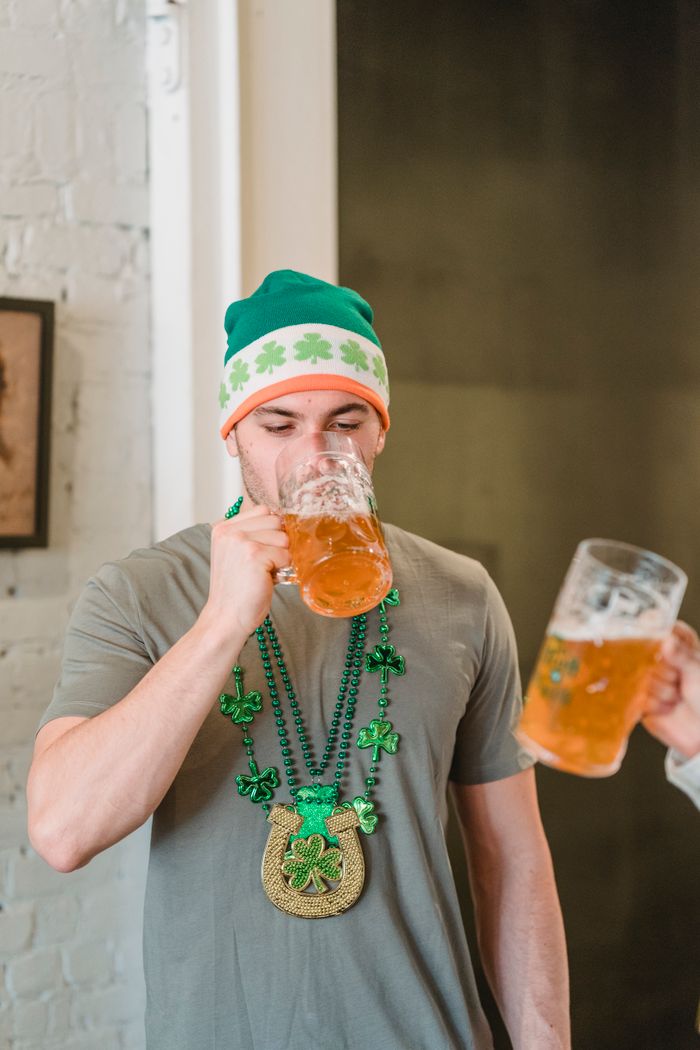 Young male in green St Patricks day decorations having glass of beer with crop anonymous friend